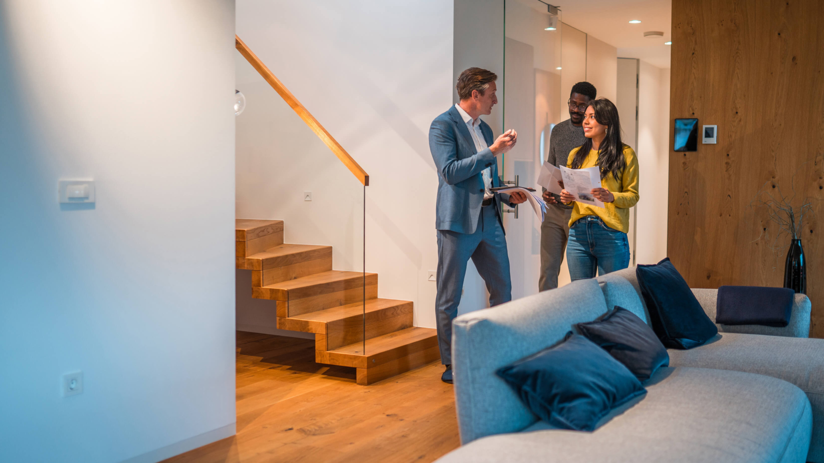 A banner image of a real estate agent guiding potential buyers through a modern home, discussing property details. The setting features a stylish interior with warm wooden accents, a cozy sofa, and elegant lighting, creating a welcoming atmosphere for decision-making.